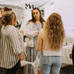 Group of brides talking with wedding vendors at a bridal fair, surrounded by wedding dresses and decor displays under string lights at the Heartland Wedding Fair.