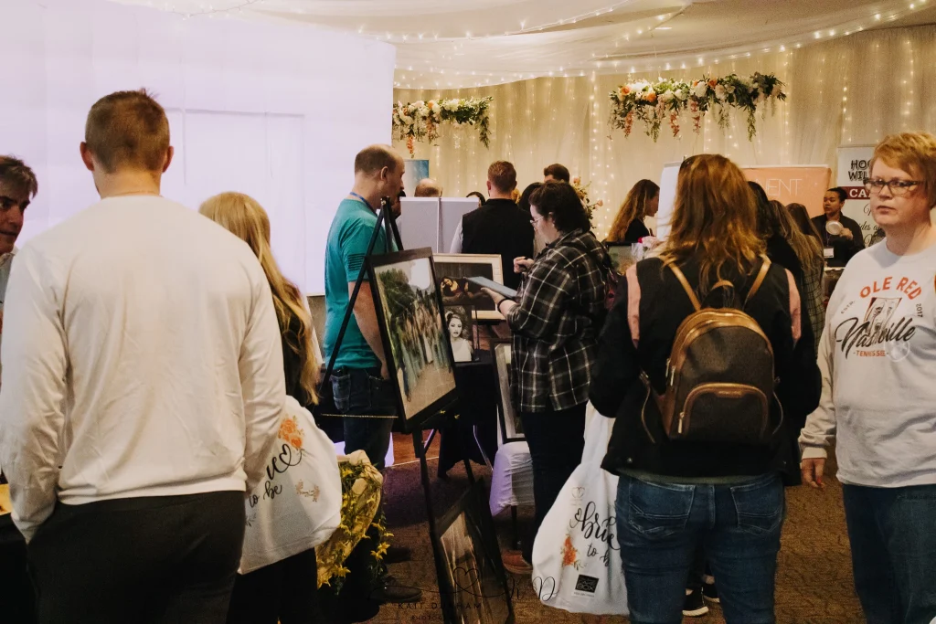 Visitors explore vendor booths and artwork displays at a bustling wedding fair and wedding expo, collecting bags of bridal goodies under twinkling lights and floral decorations.