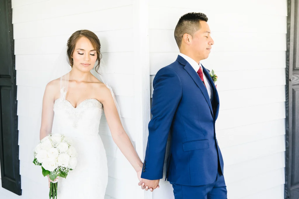 A bride and groom share a tender “first touch,” holding hands around a doorway before the ceremony, as a modern alternative to the traditional “first look,” highlighting how couples are personalizing wedding traditions for more intimate moments.